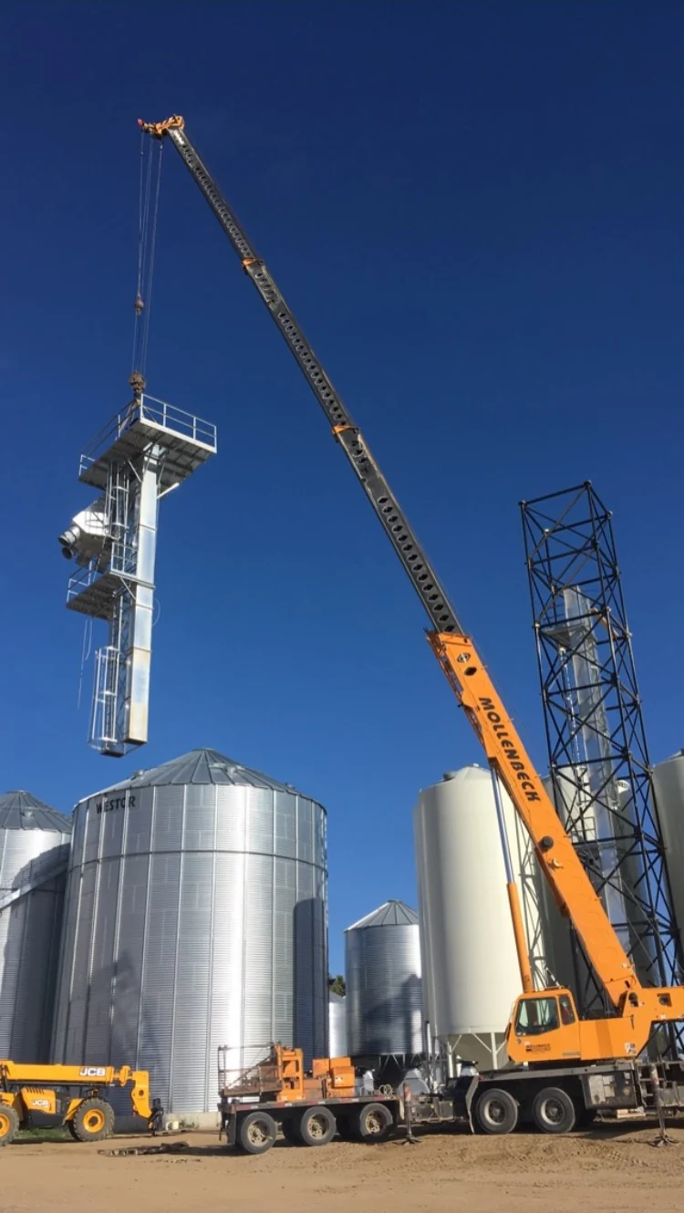 Crane lifting a bucket elevator assembly beside a steel tower and grain bins
